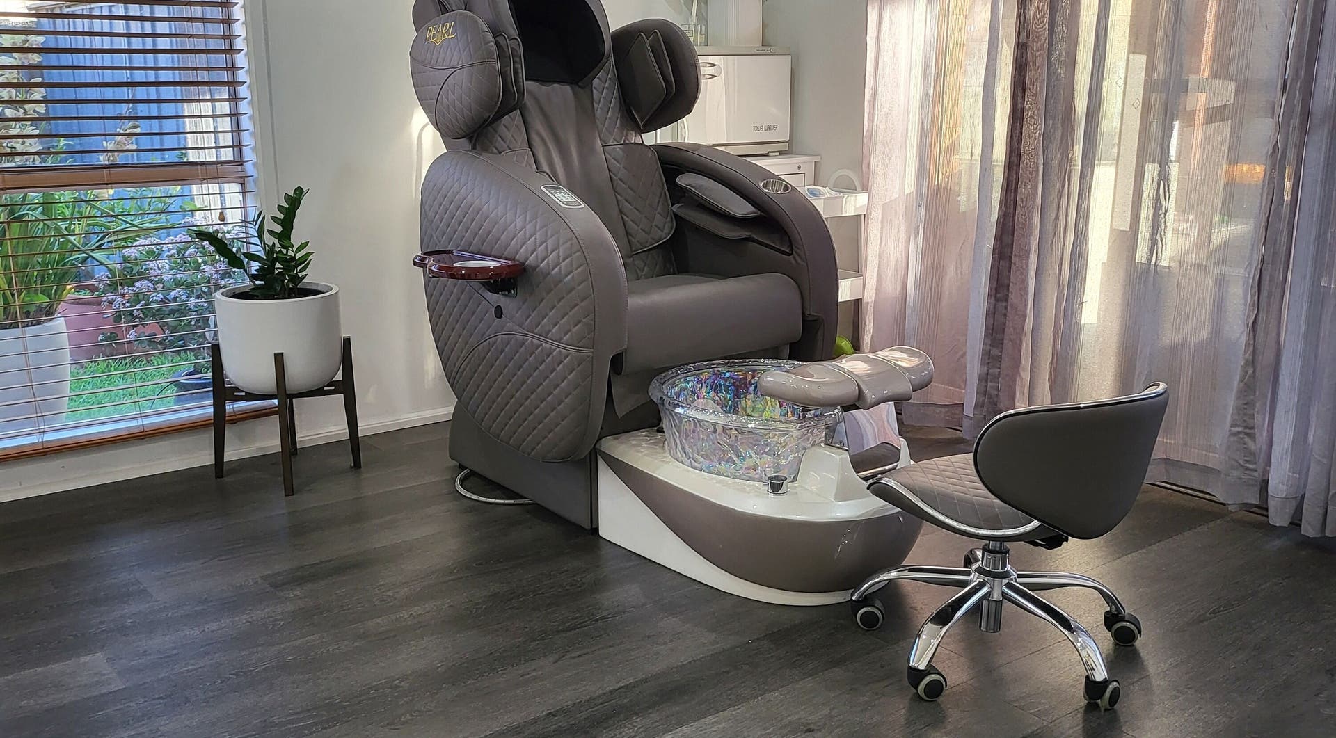 A grey quilted pedicure chair with a glass foot bath beside a sunny window